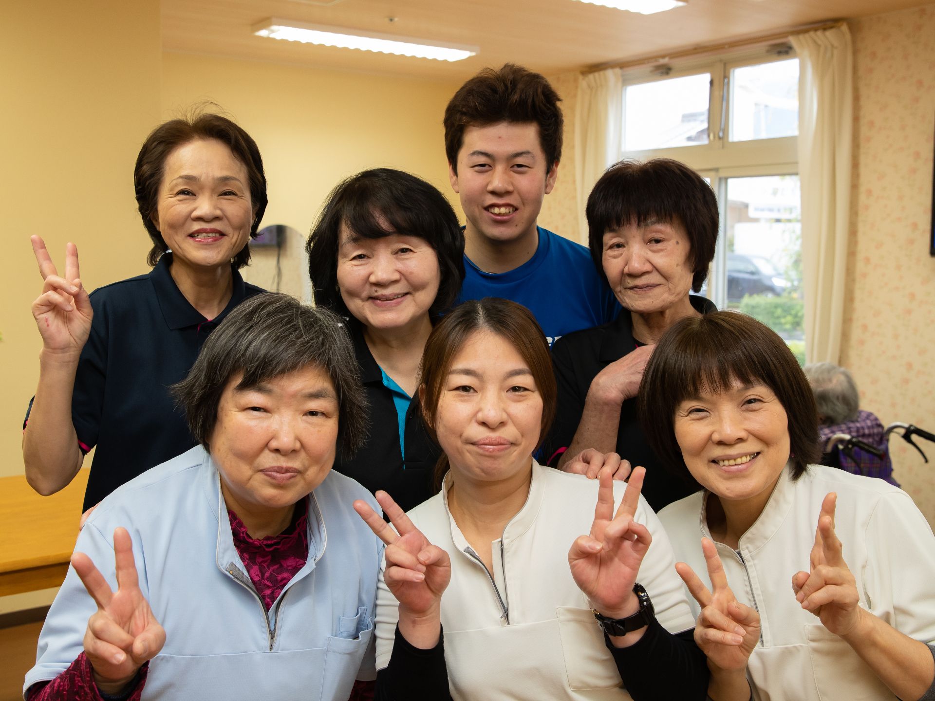 女性６人と男性１人の計７名のスタッフが２列に並んでいる。全員にこやかな顔である。右側後ろには、窓が設けられている。