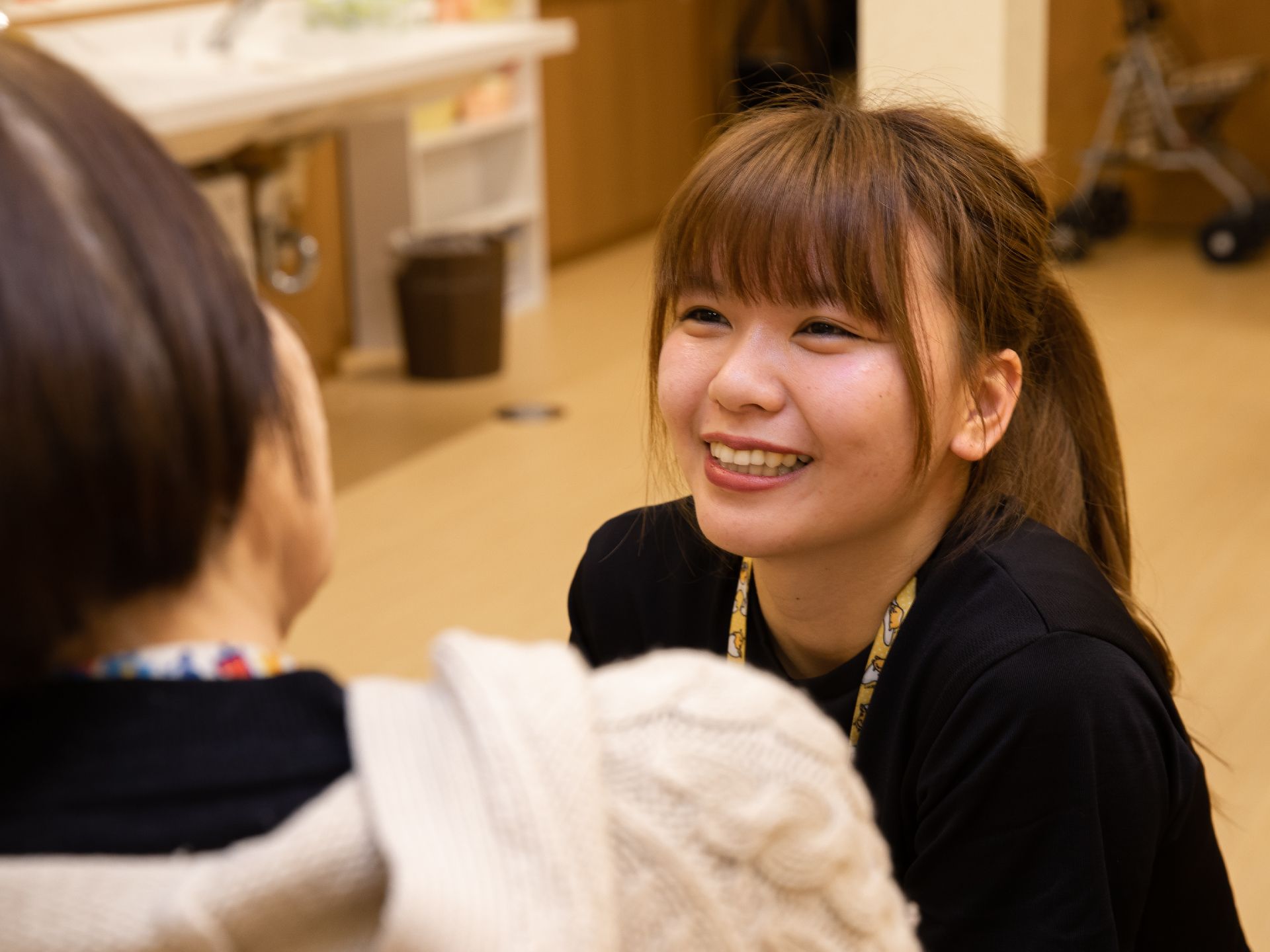 女性スタッフが明るい笑顔を入居者様に向けている。黒いポロシャツを着ている。左側の壁際に、洗面台が設置されている。