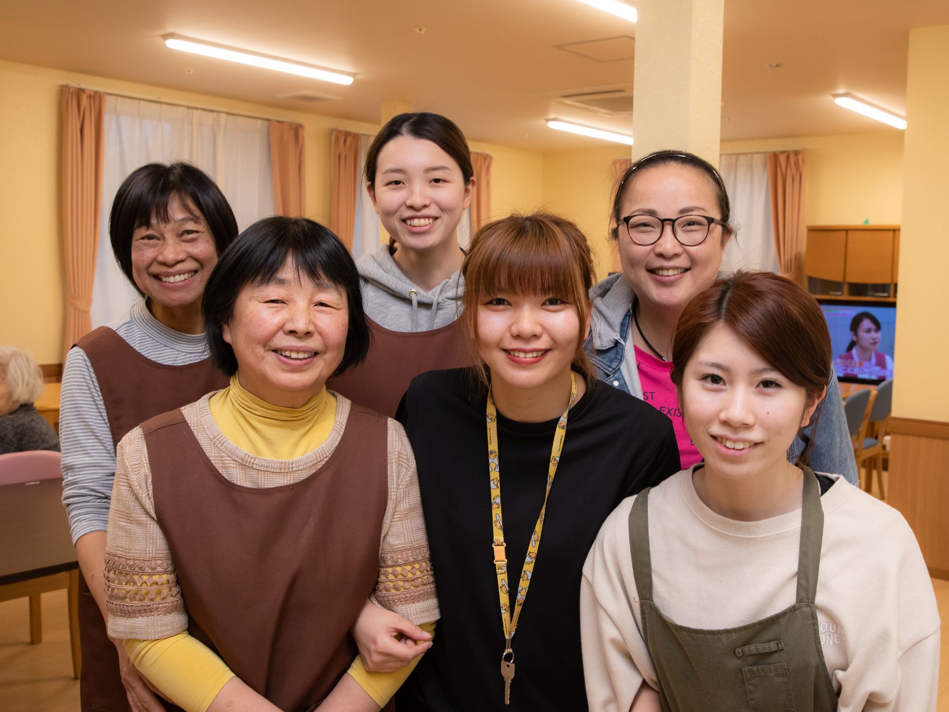 年代の違う女性スタッフが５人並んで笑顔で立っている。複数の窓が設けられており、大型テレビが右奥の壁際に設置されている。