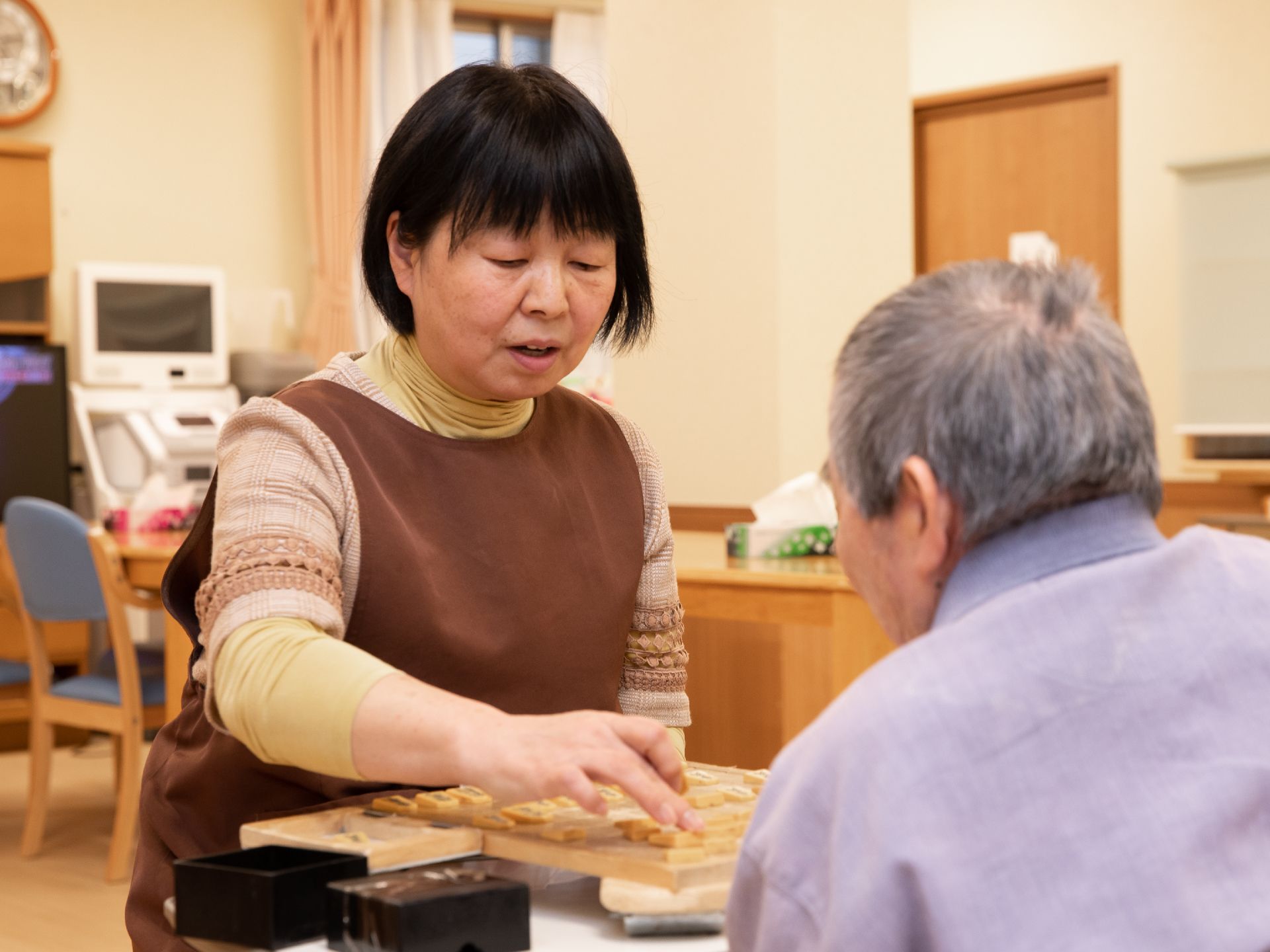 明るく温かな雰囲気の部屋で、女性スタッフが男性入居者様とテーブルで将棋を指している。時計が壁に設置されている。