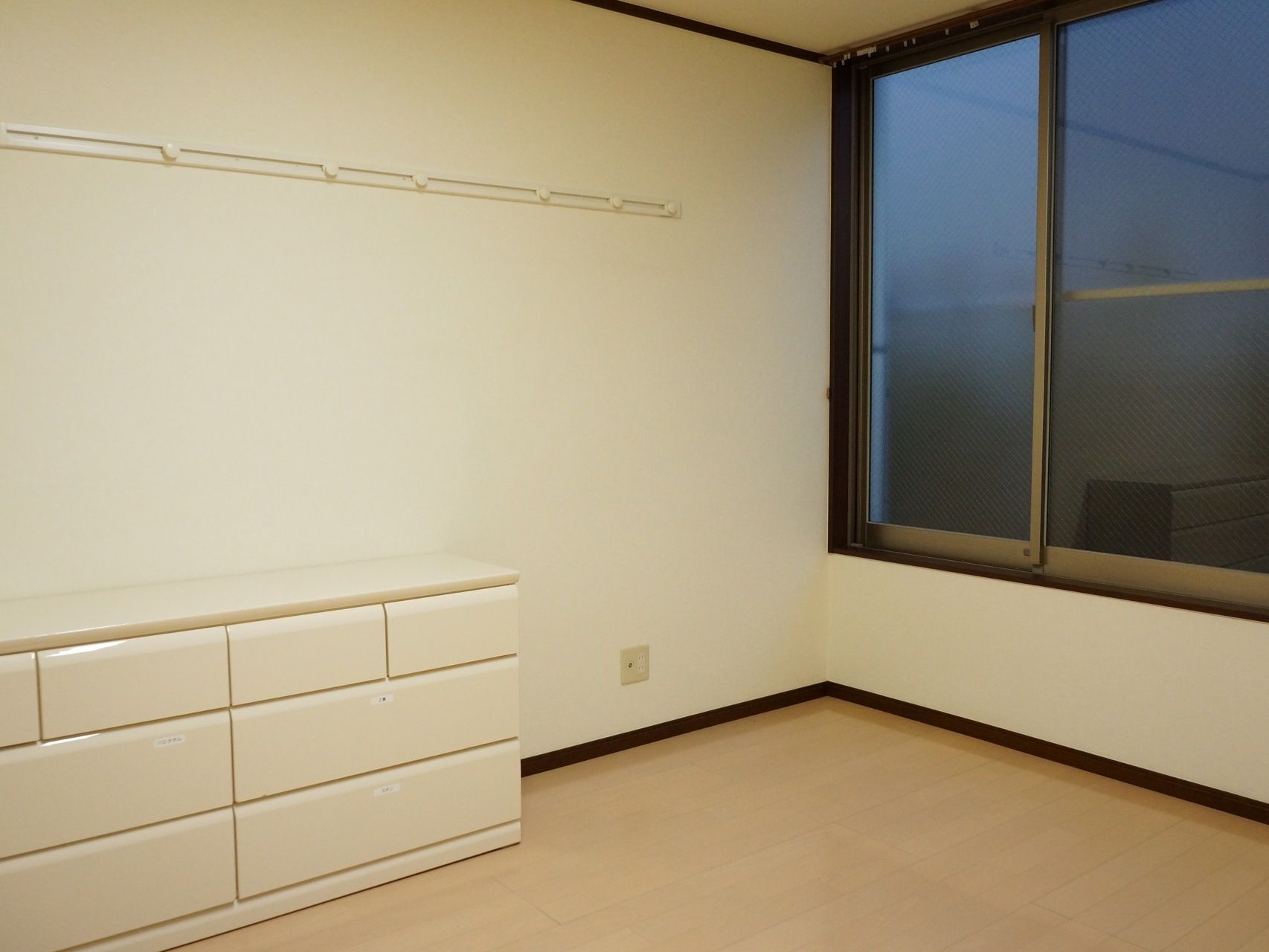 居室の床は明るいブラウンのフローリングである。壁の色は白で、左の壁際には同色のローチェストを配置している。