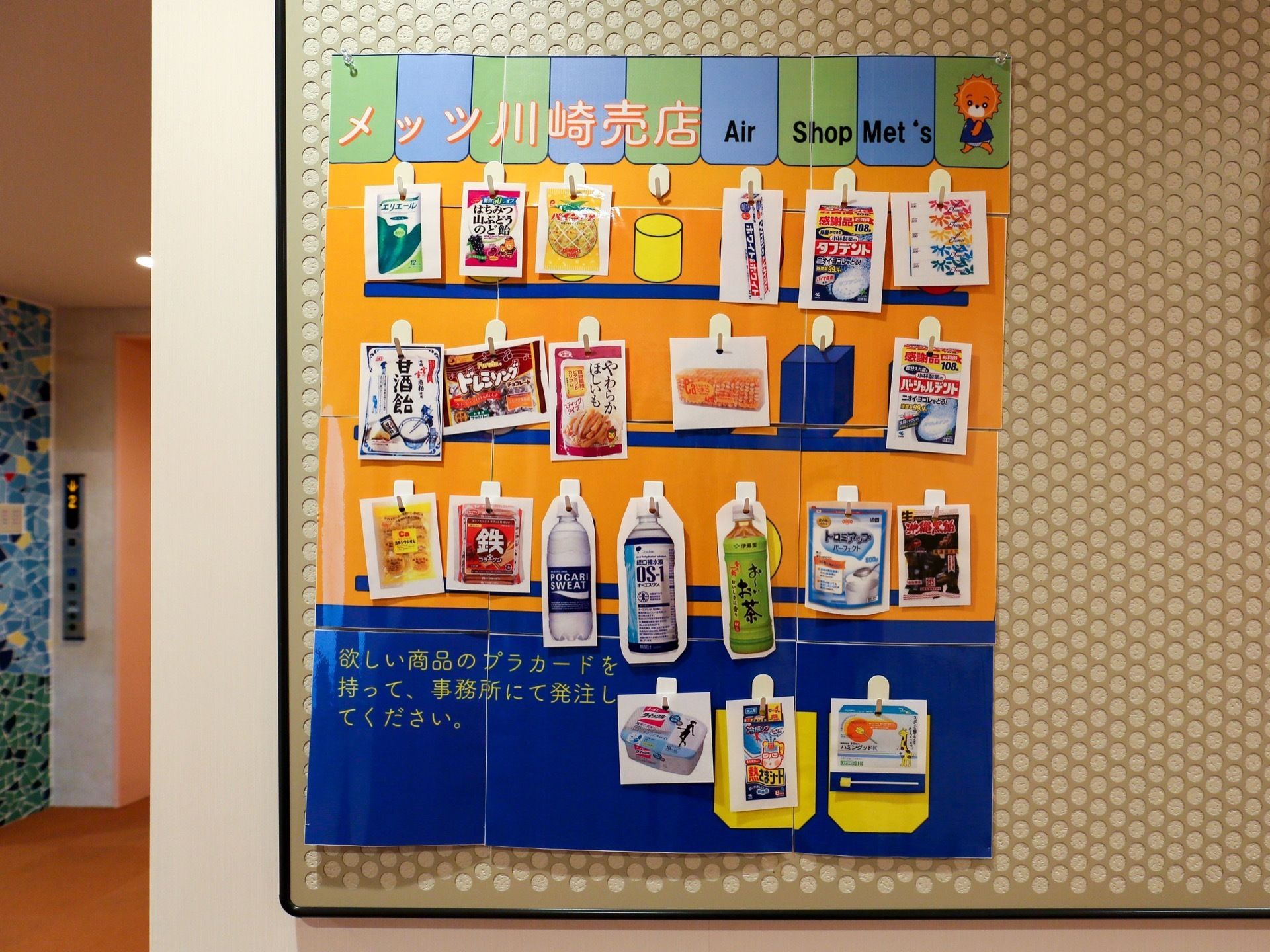 通路の壁に貼ってある、掲示物である。売店で売られている商品がひと目で分かるようになっている。奥にはエレベーターがある。