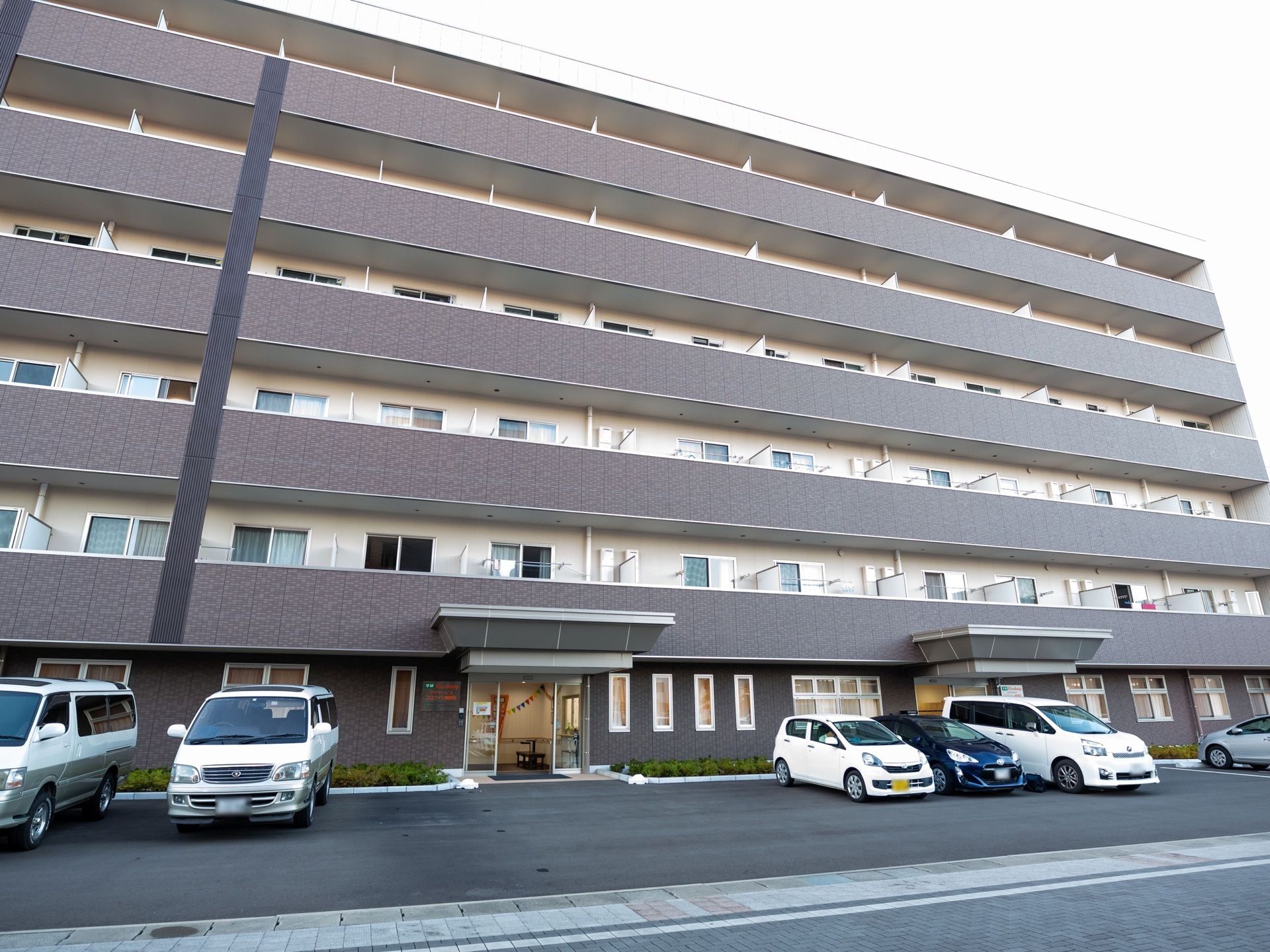 6階建てのマンションのような外観の建物は、ダークグレーと白色を基調としている。敷地内には駐車スペースが設けられている。