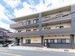 ベージュとブラウン、グレーの色合いの３階建ての外観である。１階の外壁に木造りの表札を掲げている。建物横にスロープを設けている。