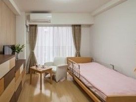 レースのカーテンと遮光カーテンが掛かった掃き出し窓がある。壁面には引き出し家具、ベッドの横には一人掛けソファとテーブルが置かれている。