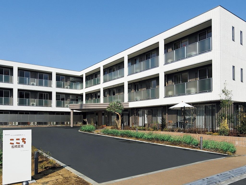 白色の大きなマンションのような外観。駐車場の前には施設名の書かれた看板が立てられている。各部屋にバルコニーがある。