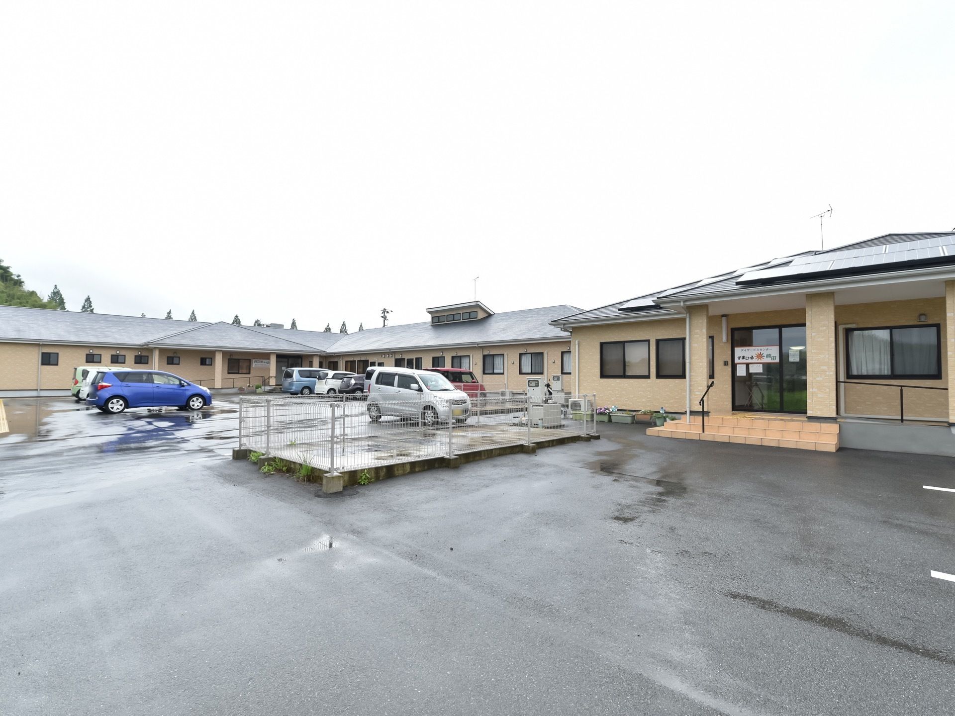 施設の屋根には、ソーラーパネルやテレビのアンテナが設置されている。建物の前には駐車場があり、車が数台停まっている。