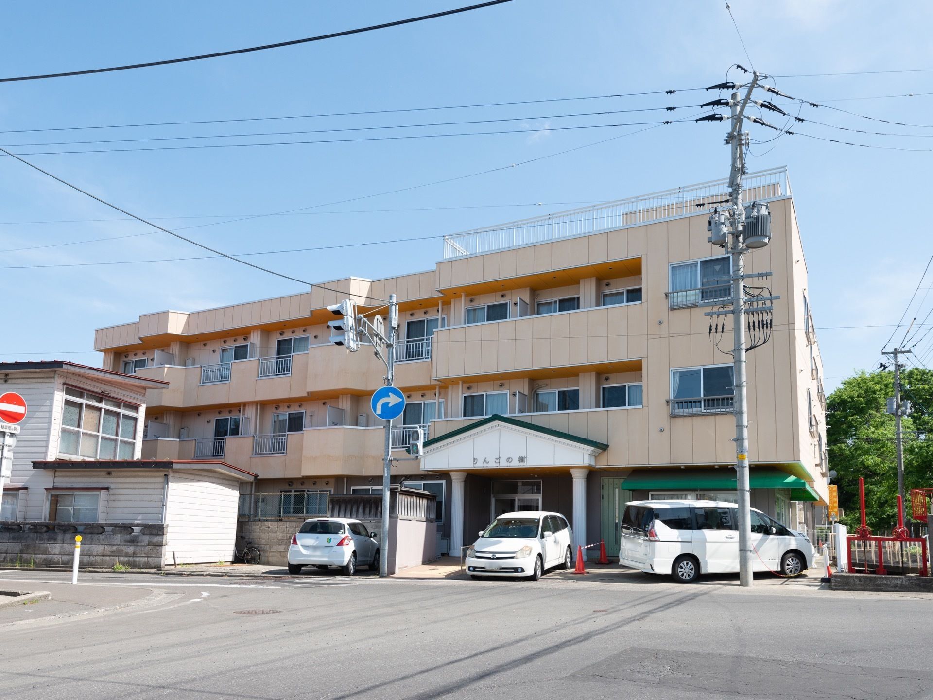 車道に面した敷地に淡いオレンジ色の3階建ての建物が建っている。玄関の近くに白い車が２台停められている。