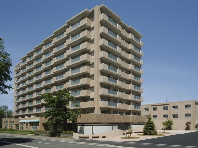 ベージュ外壁のマンションのような大きな建物は落ち着いた雰囲気で、各フロアにベランダがあり、周辺には木が植えられている。