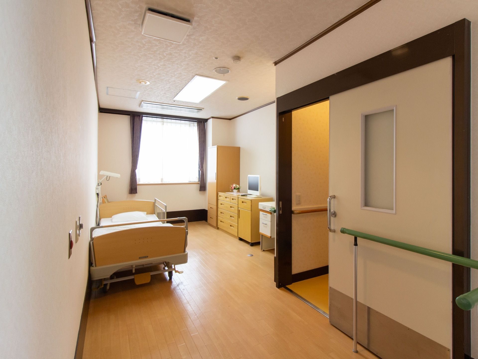 奥行きのある部屋に、ベッドや収納家具が置かれている。トイレの左右に緑色の手すりが施されており、扉は引き戸になっている。