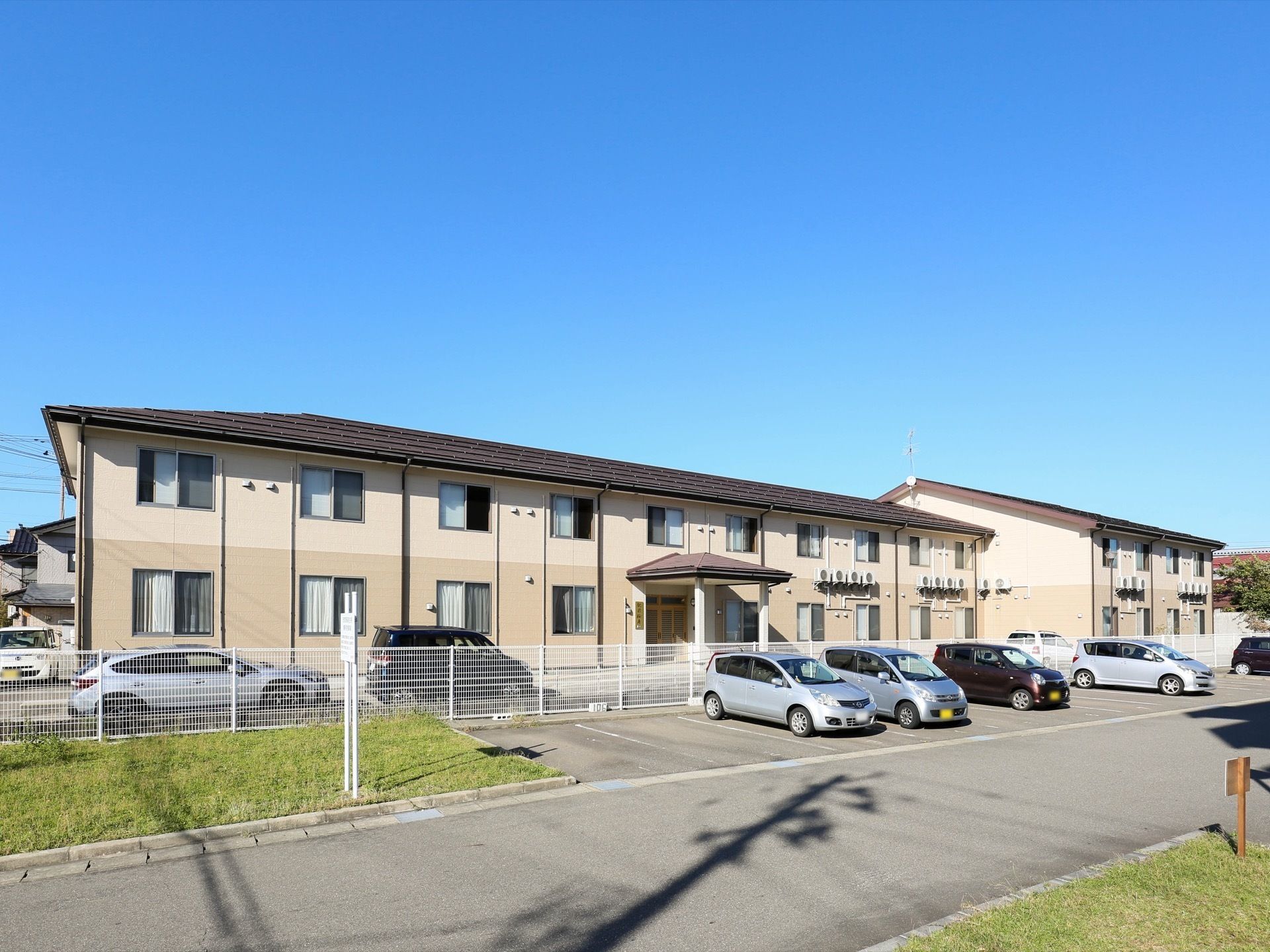 ２階建ての明るい色合いの施設が建てられている。敷地内には、駐車スペースが多数設けられている。周辺には住宅が見える。