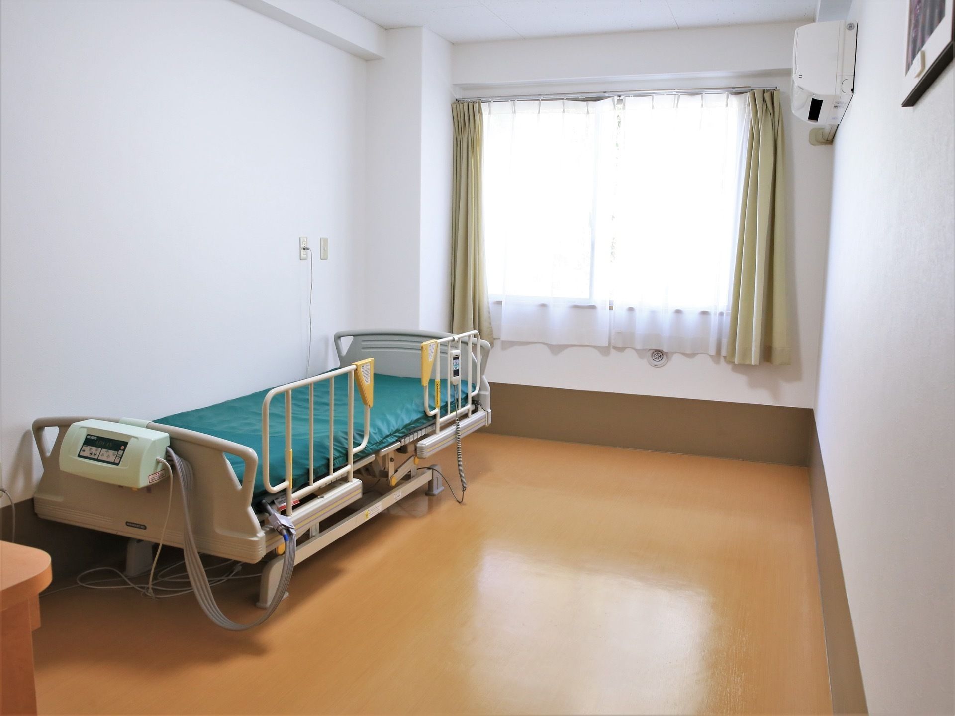 居室には大きな窓があり、カーテンが取り付けられている。部屋にはエアコンや介護用ベッドが設置されている。