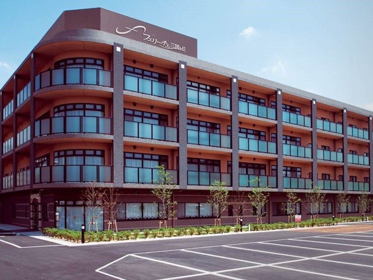 大きな駐車場が付いた4階建ての建物はブラウン系の外壁で、建物の周りには草木が植えられている。