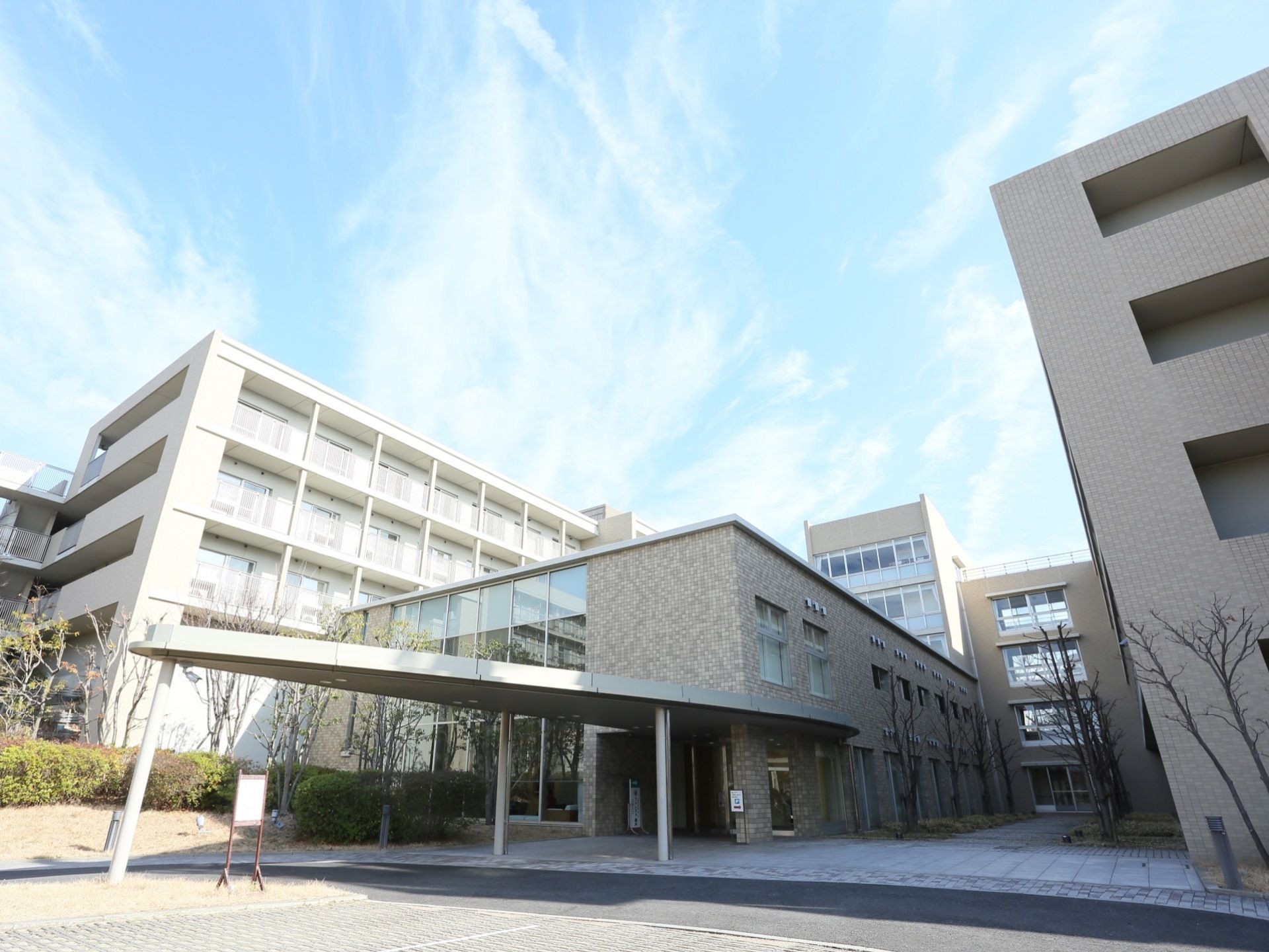 大型の施設は、グレーの外壁と白の外壁の建物が入り組む構造である。手前や中庭には、多数の樹木が植えられている。