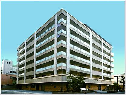 シルバーのような色の外観の大きな建物の角フロアにベランダがあり、周りにはたくさん建物がある。