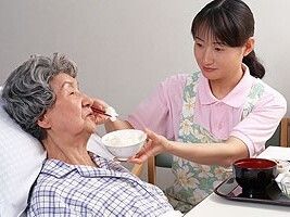 エプロンを付けた女性スタッフがベッドにいるパジャマを着た高齢女性に白いご飯を食べさせている。