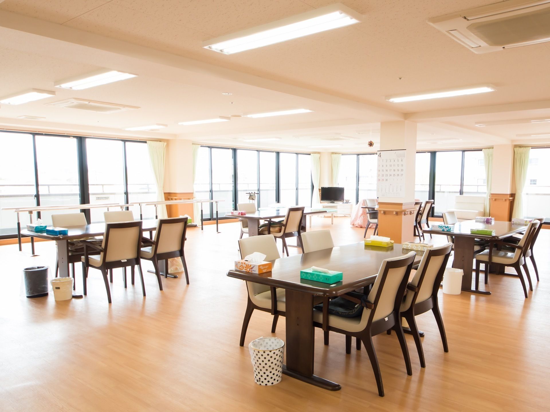 平行棒やテレビが設置されている、食堂兼機能訓練室である。各テーブルの上には箱ティッシュが用意されている。