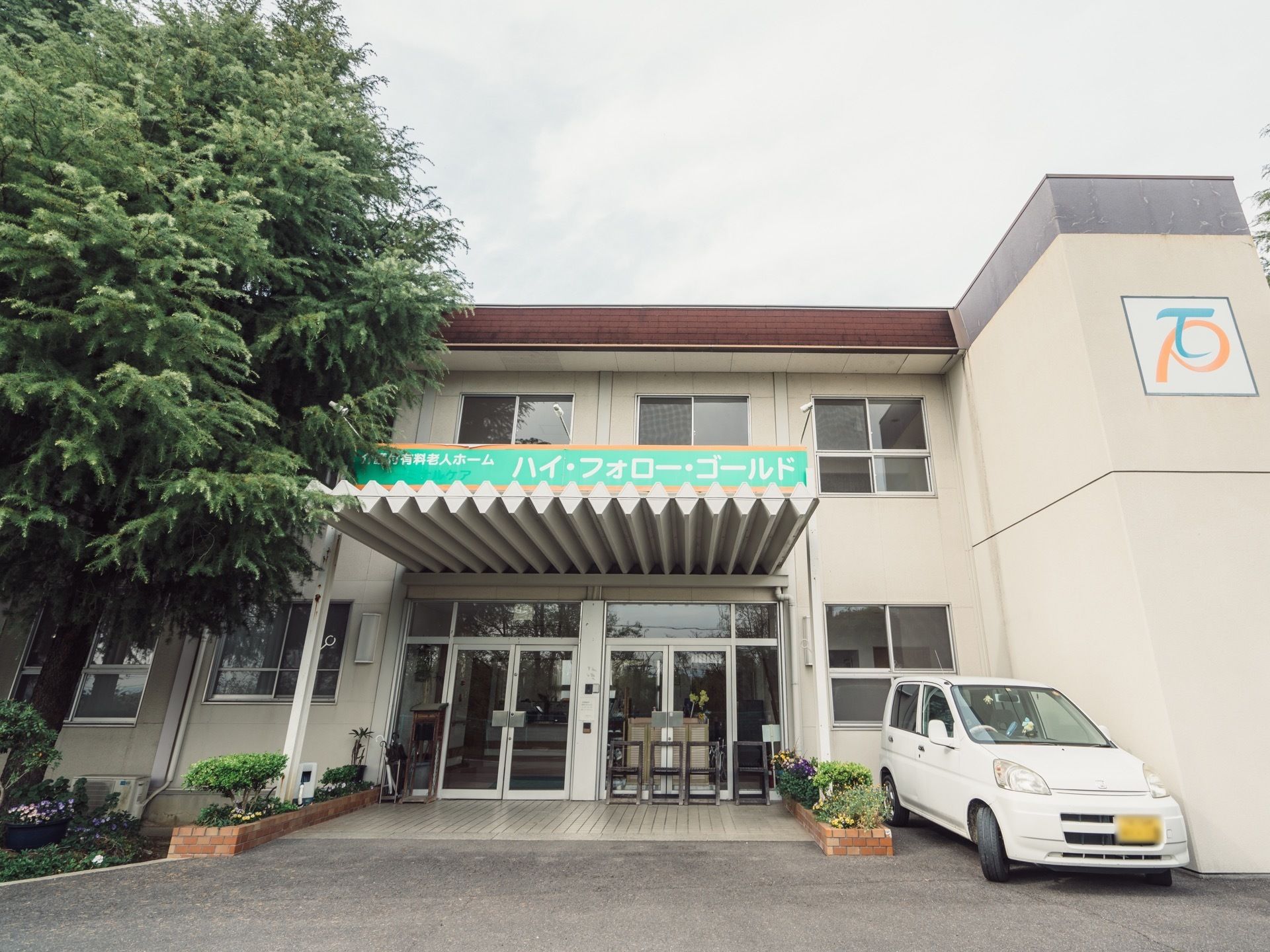 波型の小屋根のある玄関である。玄関上には、施設名が書かれた看板がつけられ、横には車が１台停まっている。