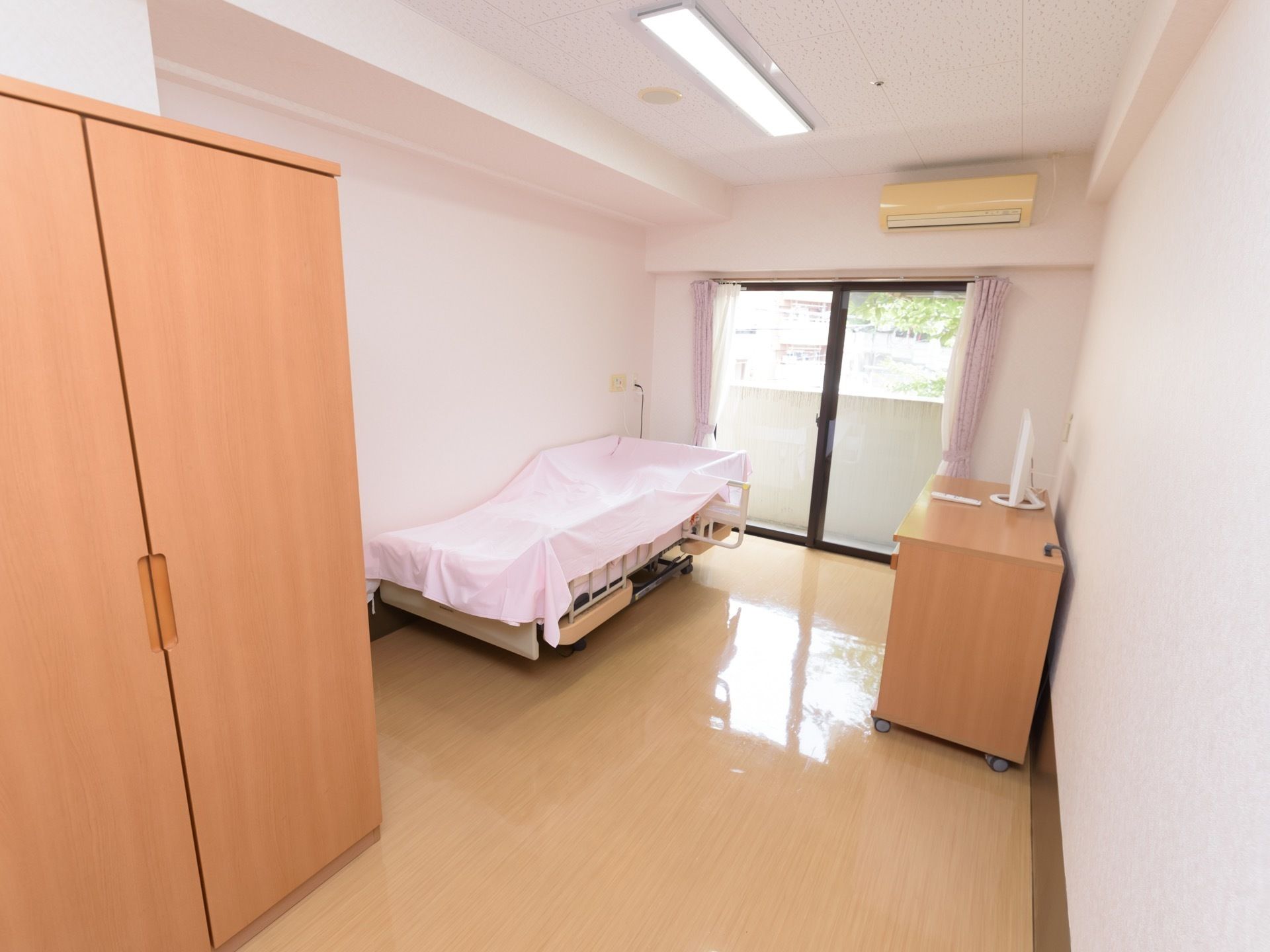 居室の窓は掃き出し窓で、室内が明るい。柵が付いた介護用ベッド、エアコン、キャスター付きのローボードがある。