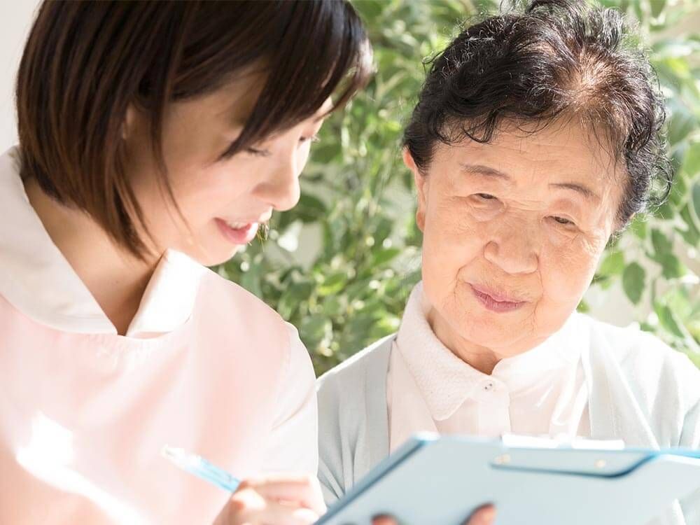 カルテのようなものを見ながら話しているエプロンを付けた若い女性スタッフとカーディガンを着た高齢女性。