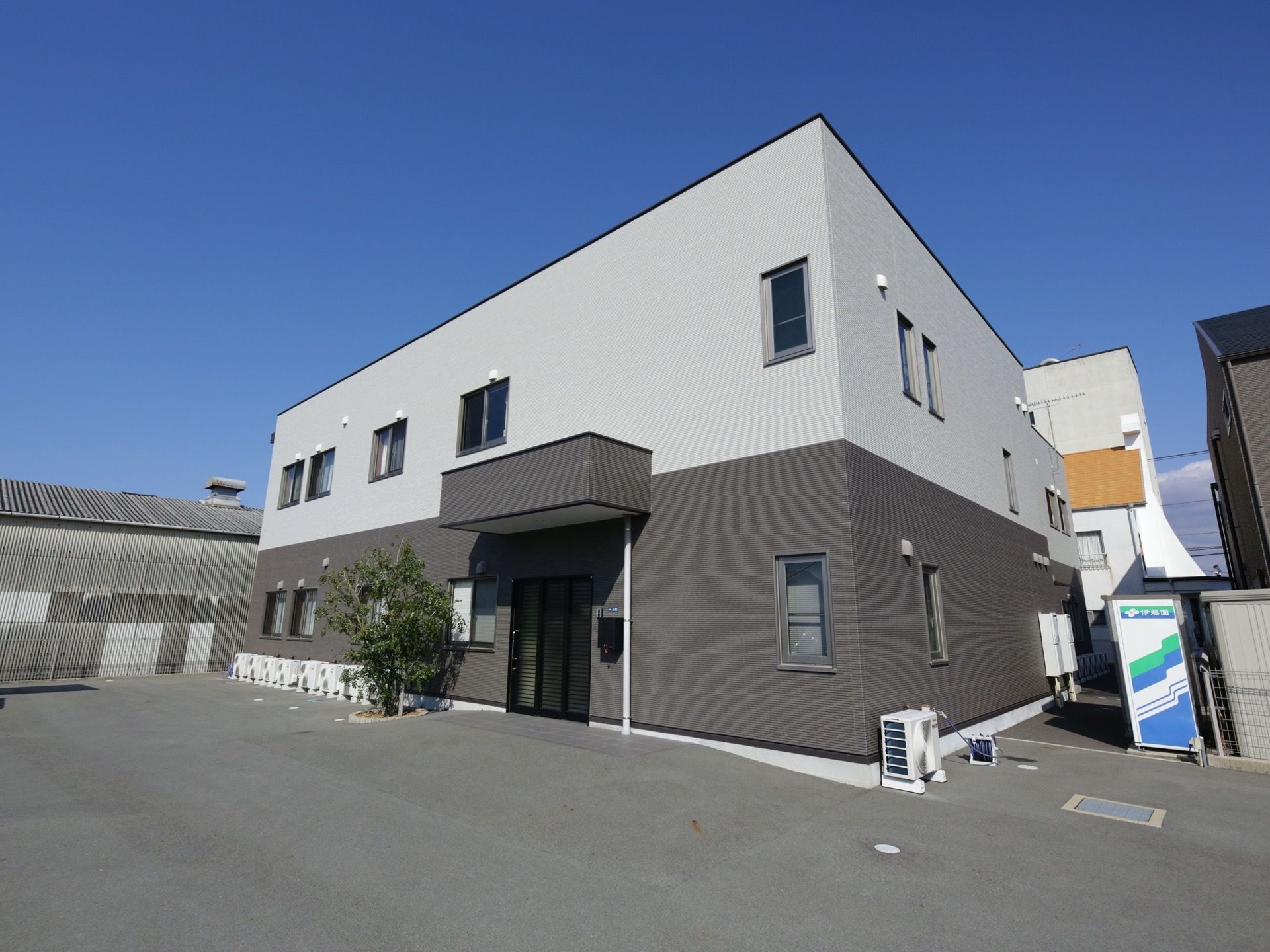 建物の横に自動販売機が１台置かれており、敷地内はアスファルトで舗装されている。玄関には、小さな屋根が設置されている。