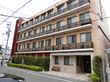 ベージュ系の4階建ての建物。各フロアにベランダが付き、建物の周りには緑が植えられ、玄関にベンチもある。