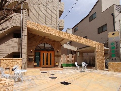 「応援家族 成増」の外観。高いマンション街に位置する、5階建てのベージュ色の建物。