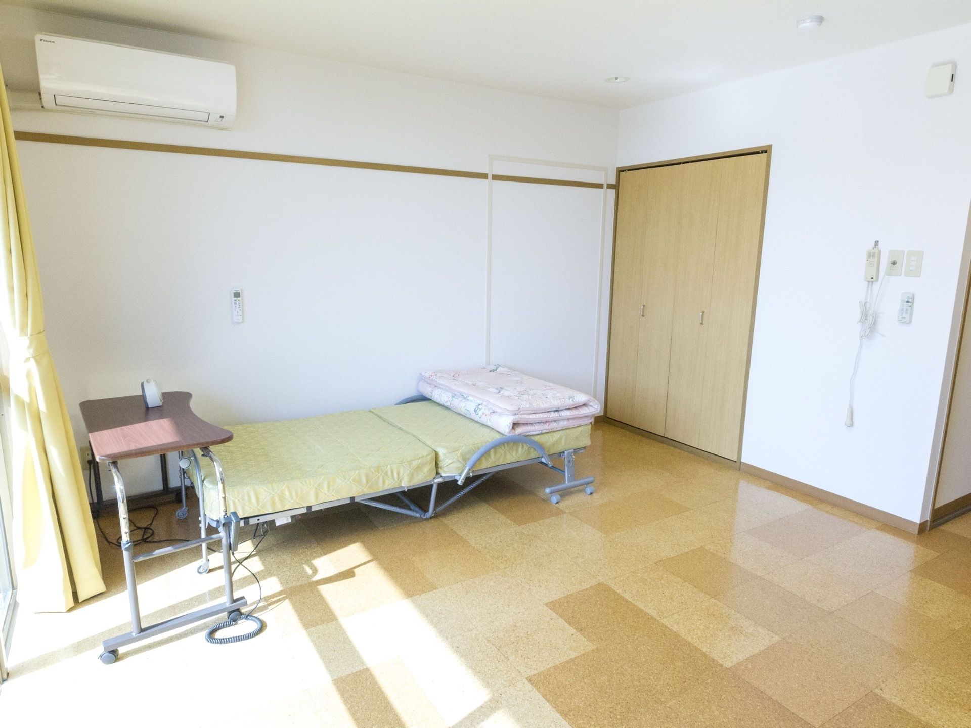 壁にエアコンが設置されている。木目調の扉のついた収納がある。ベッドの横の壁にはリモコンが掛けられている。