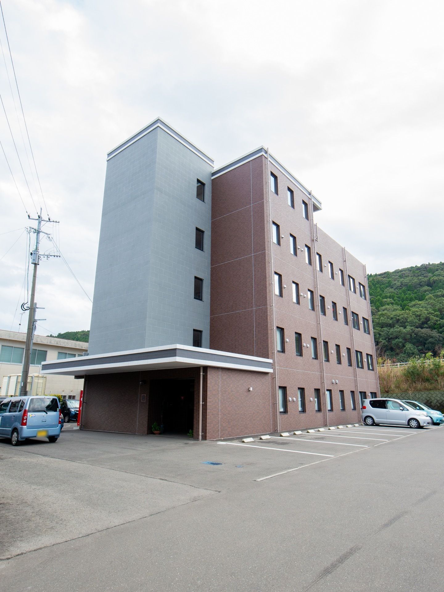広い駐車場のある５階建ての建物で、裏には森がある。玄関には屋根があり、奥の壁際に自動販売機が置かれている。