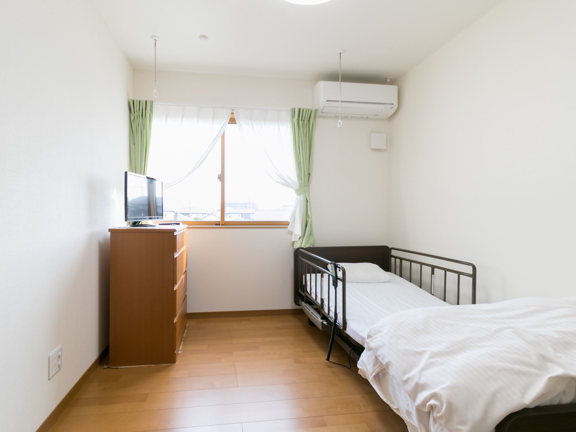 個室の居室である。太陽光が差し込む窓が設けられている。介護ベッドやタンスが置かれている。壁には、エアコンが設置されている。
