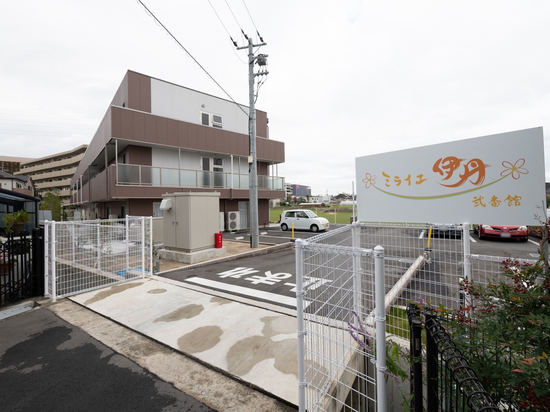 建物の傍らに広い駐車場がある。フェンスで囲まれ、舗装されている。入口付近には施設の名前が書かれた大きな看板が設置されている。