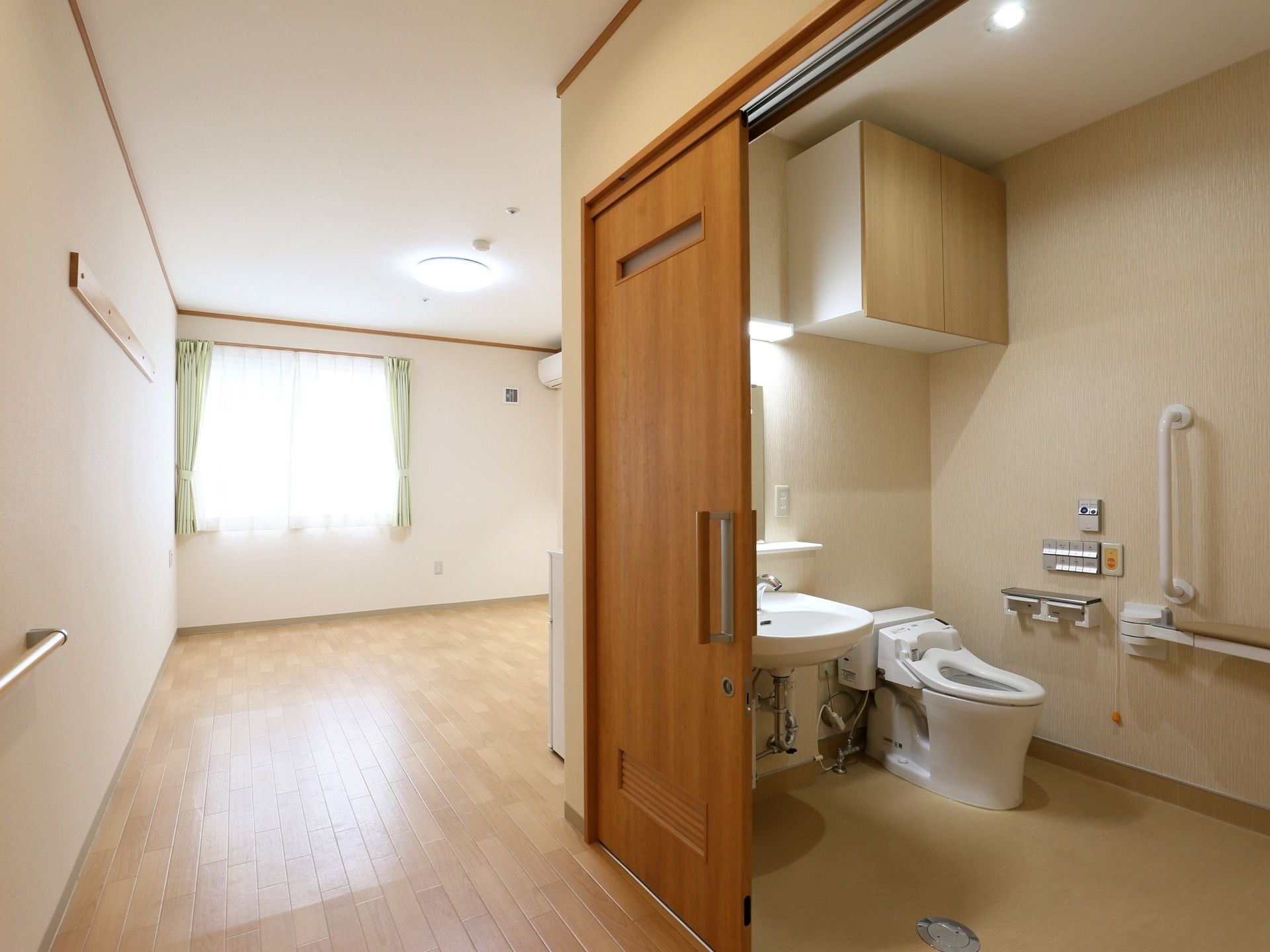 居室に入ると右手に車椅子対応の広いトイレがある。床はフローリングで、突き当りには窓があり、カーテンが掛かっている。