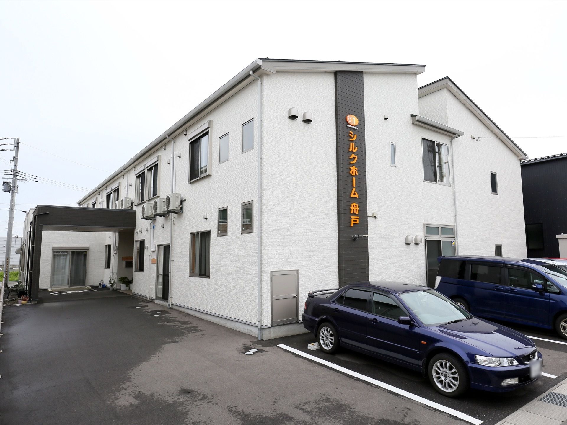 白い外壁の大きな2階建ての建物があり、その前に車が数台止められている。建物の入り口は屋根があり、車が寄せられる。