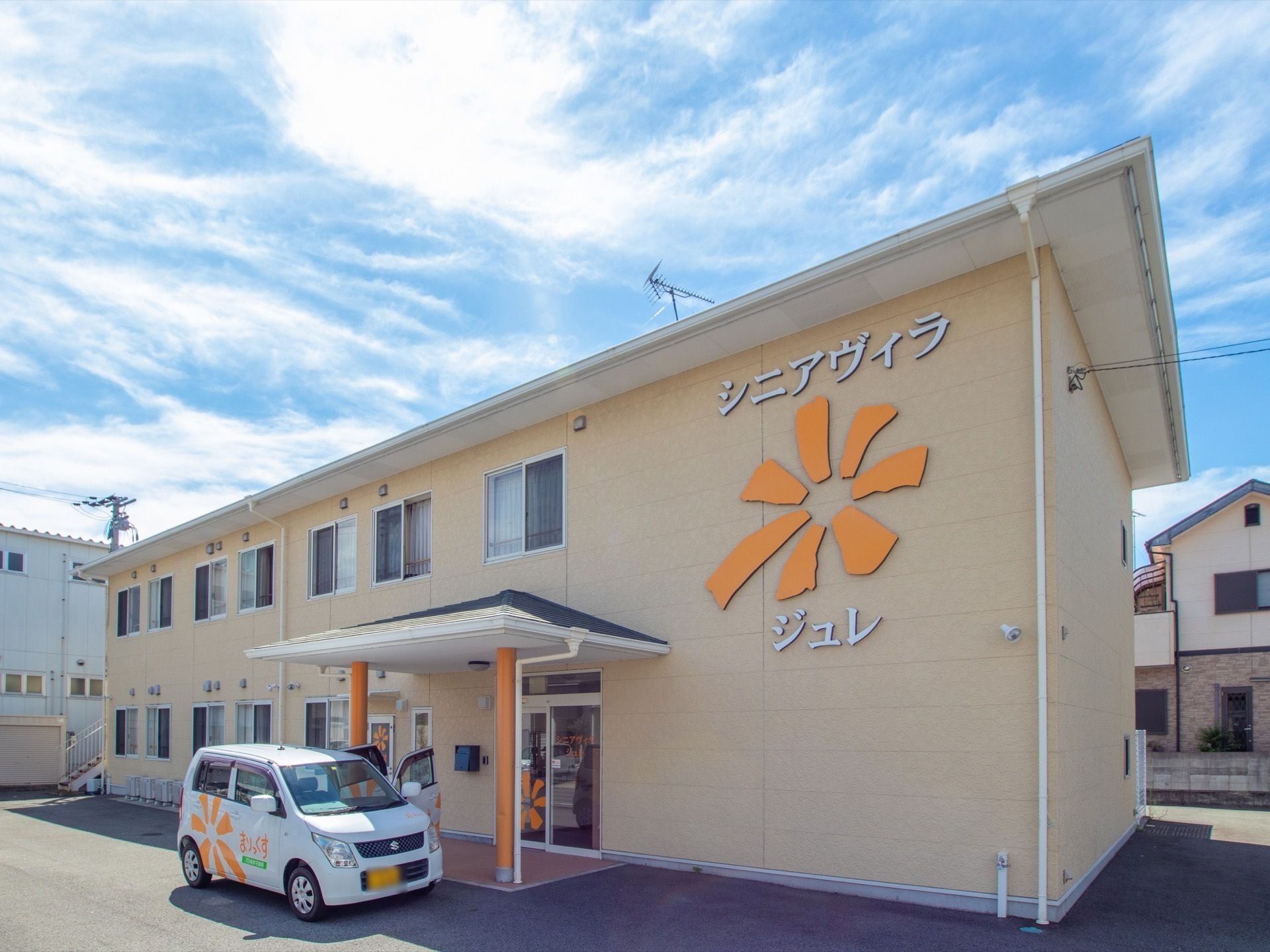 施設はベージュ色の外壁で２階建ての建物である。壁に大きく施設名称とロゴが掲示されている。玄関前に施設車両が停まっている。