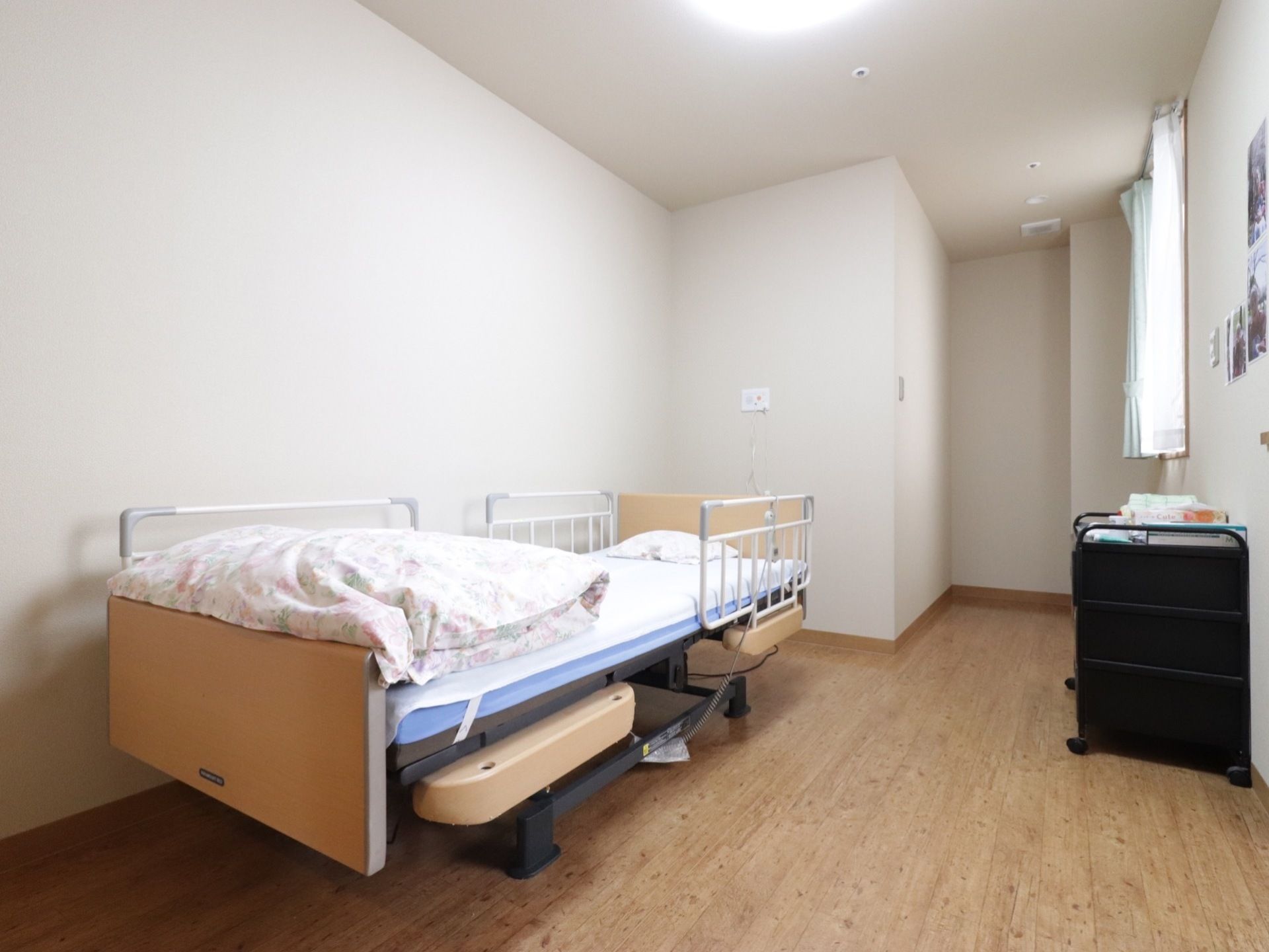 介護用ベッドが置かれている。腰高窓があり、レースのカーテンと緑のカーテンが掛けられている。床はフローリングである。