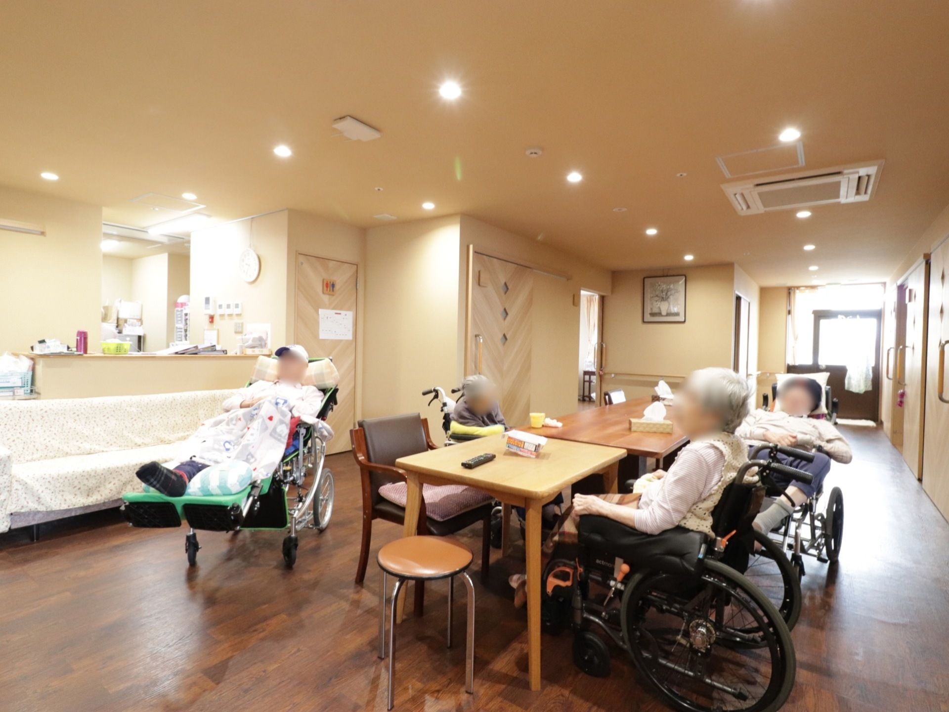 テーブルを囲んで入居者様が数人いらっしゃる。テーブルの上にティッシュボックスやリモコンが置かれている。共有トイレがある。