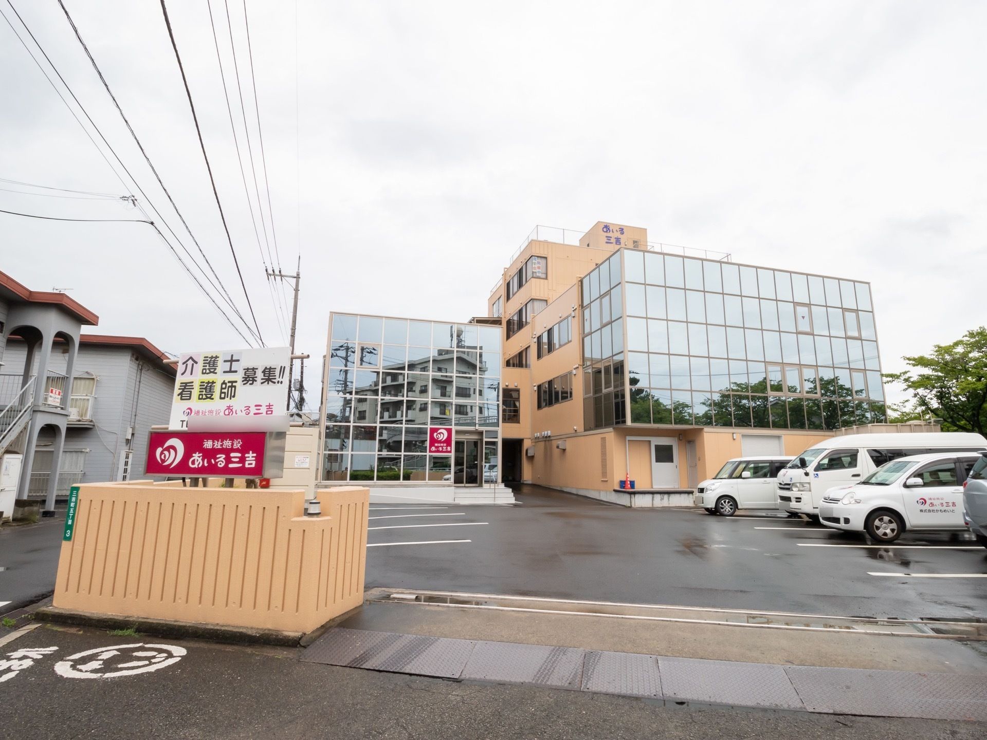 外壁の一部がミラーガラスで覆われていて、周辺の建物や樹木が写っている。手前の駐車場には、社用車も停めてある。