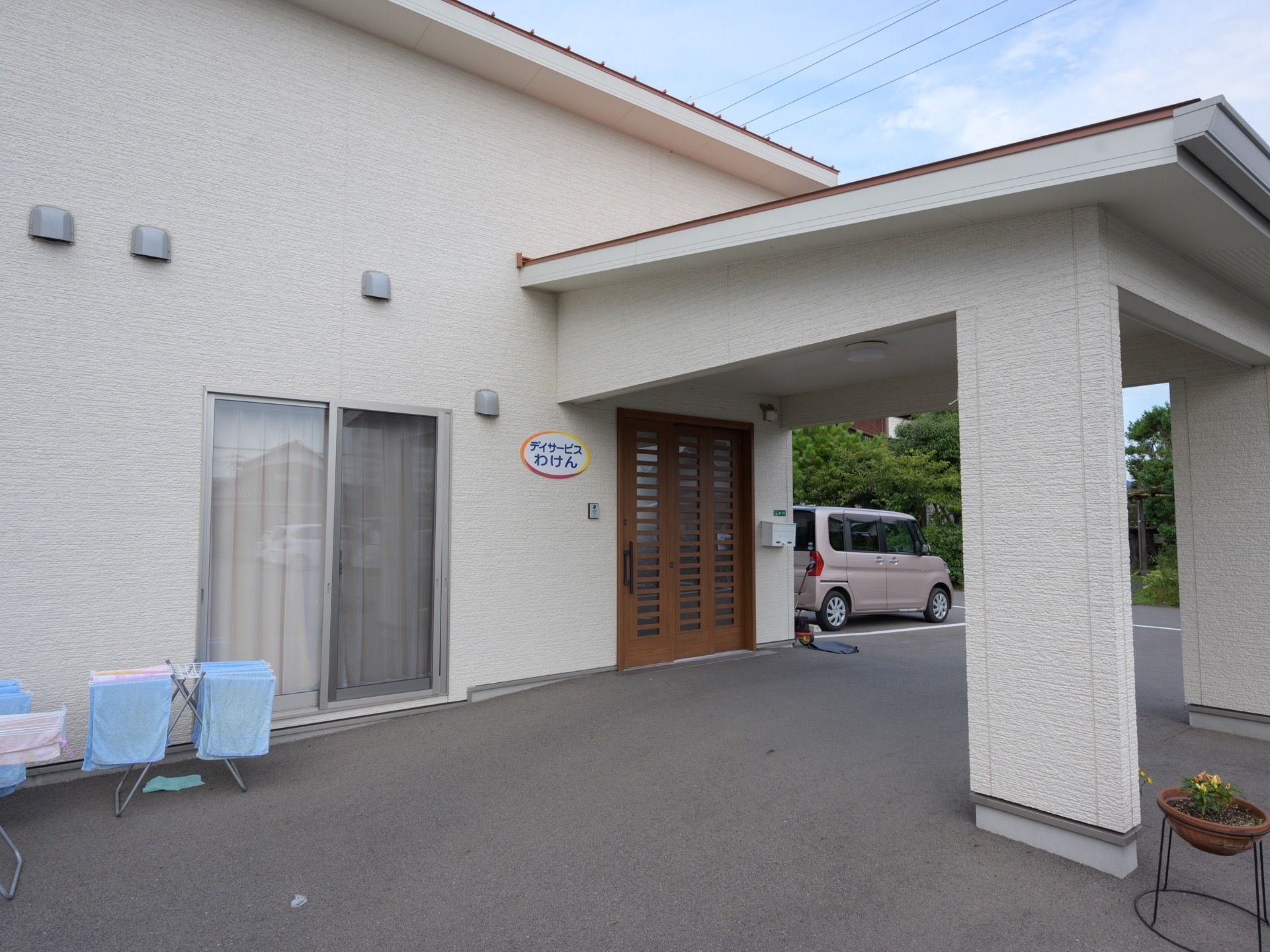 施設の建物は白色で、茶色の玄関のドアの前には車寄せが設けられている。プランターに花が植えられ、玄関前に置かれている。