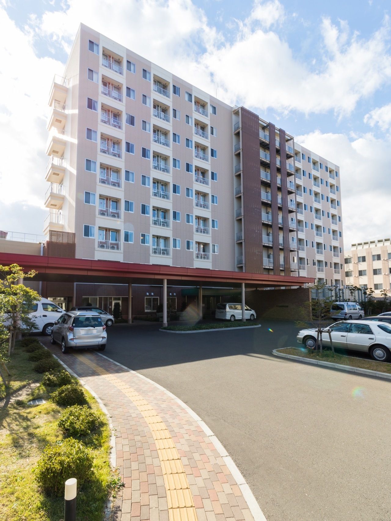 ９階建てのアイボリーの建物。歩道にはレンガが敷かれ、建物と同系色でまとめられている。周囲には緑が植えられ、手前は駐車場になっている。