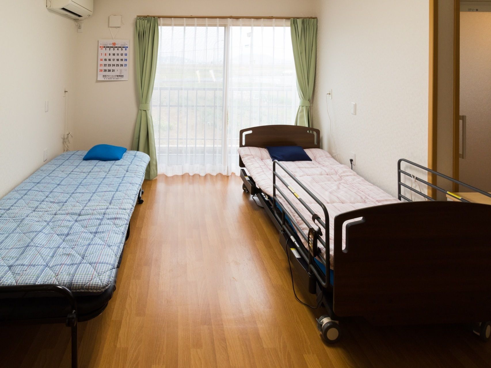 居室には大きめの窓、カーテン、壁にはエアコン、カレンダーがあり、電動介護ベッドと補助ベッドが置かれている。