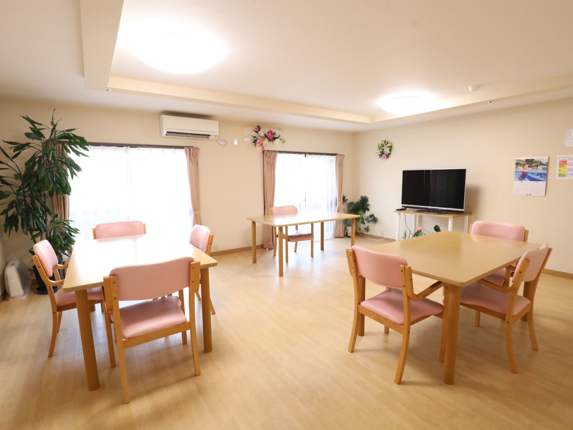 観葉植物が部屋の隅に置いてあり、壁には花束が飾られている。テレビの近くには、カレンダーが貼ってある。