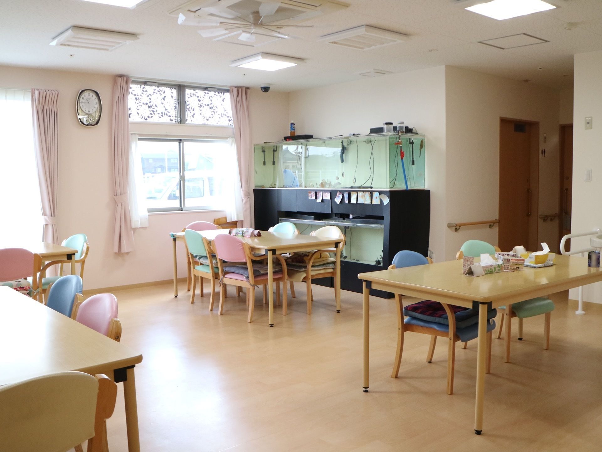 窓の淡いピンク色のカーテンが備え付けられた、共有スペースである。壁側には、大きな水槽が設置されている。