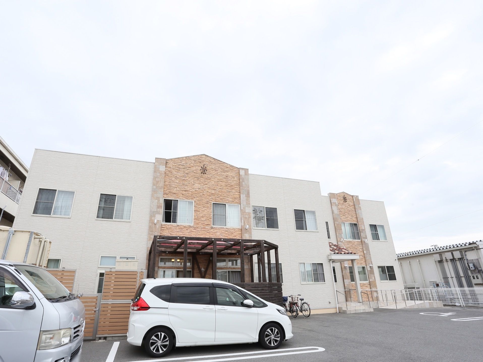 施設は2階建ての建物で、白とレンガ調の外壁になっている。建物の前には駐車場があり、車が2台停まっている。