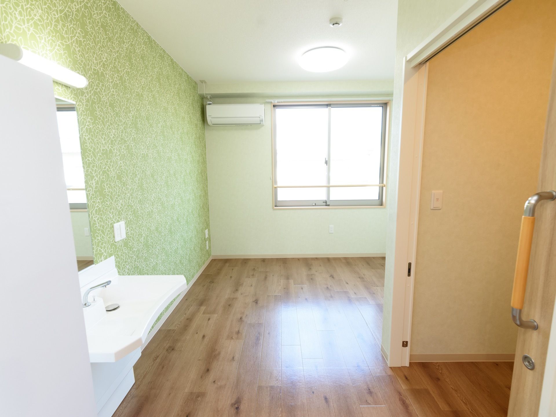 木目のフローリングの床のナチュラルな雰囲気の部屋である。腰高窓に手すりが設置され、天井の照明が点灯している。