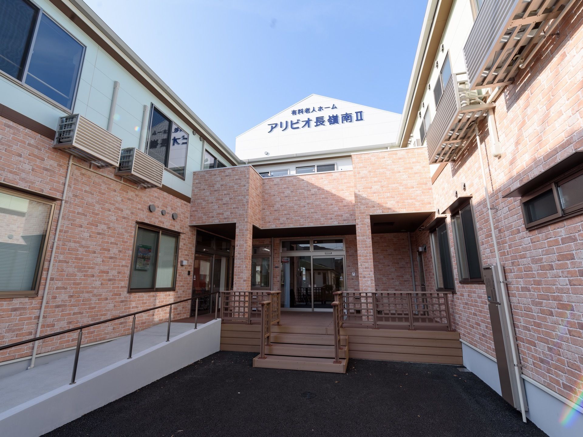 コの字型に建てられた２階建ての建物の２階の白い外壁に青い文字で施設の名称が表示されている。手すりが設置されたスロープがある。