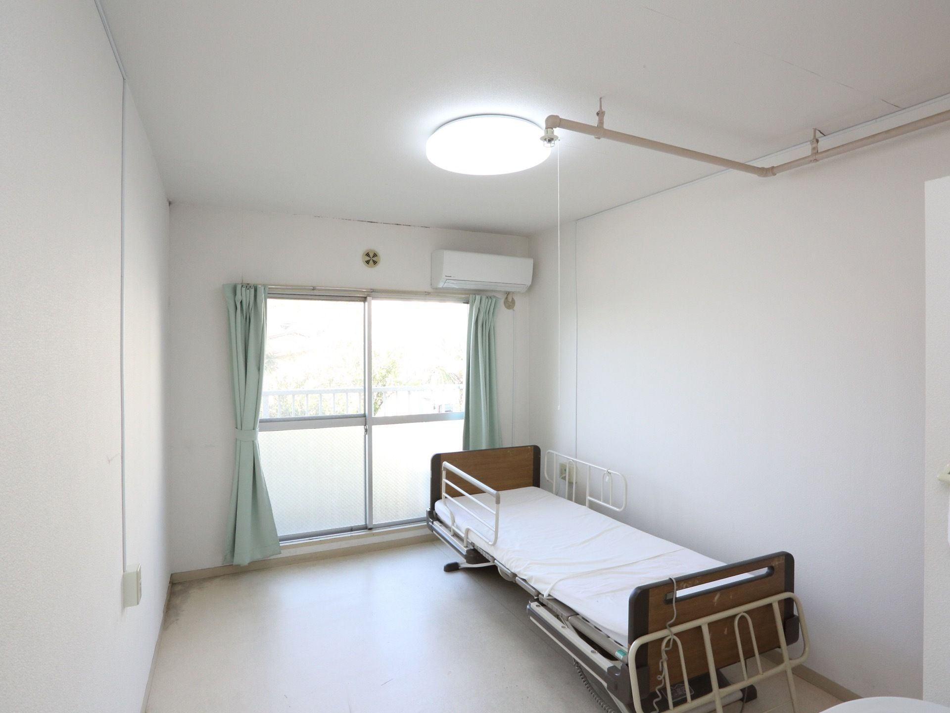 清潔感のある白い壁に囲まれた入居者様の居室。介護用ベッドが置かれ、大きな窓があり日当たりが良い。窓の外にはベランダがある。