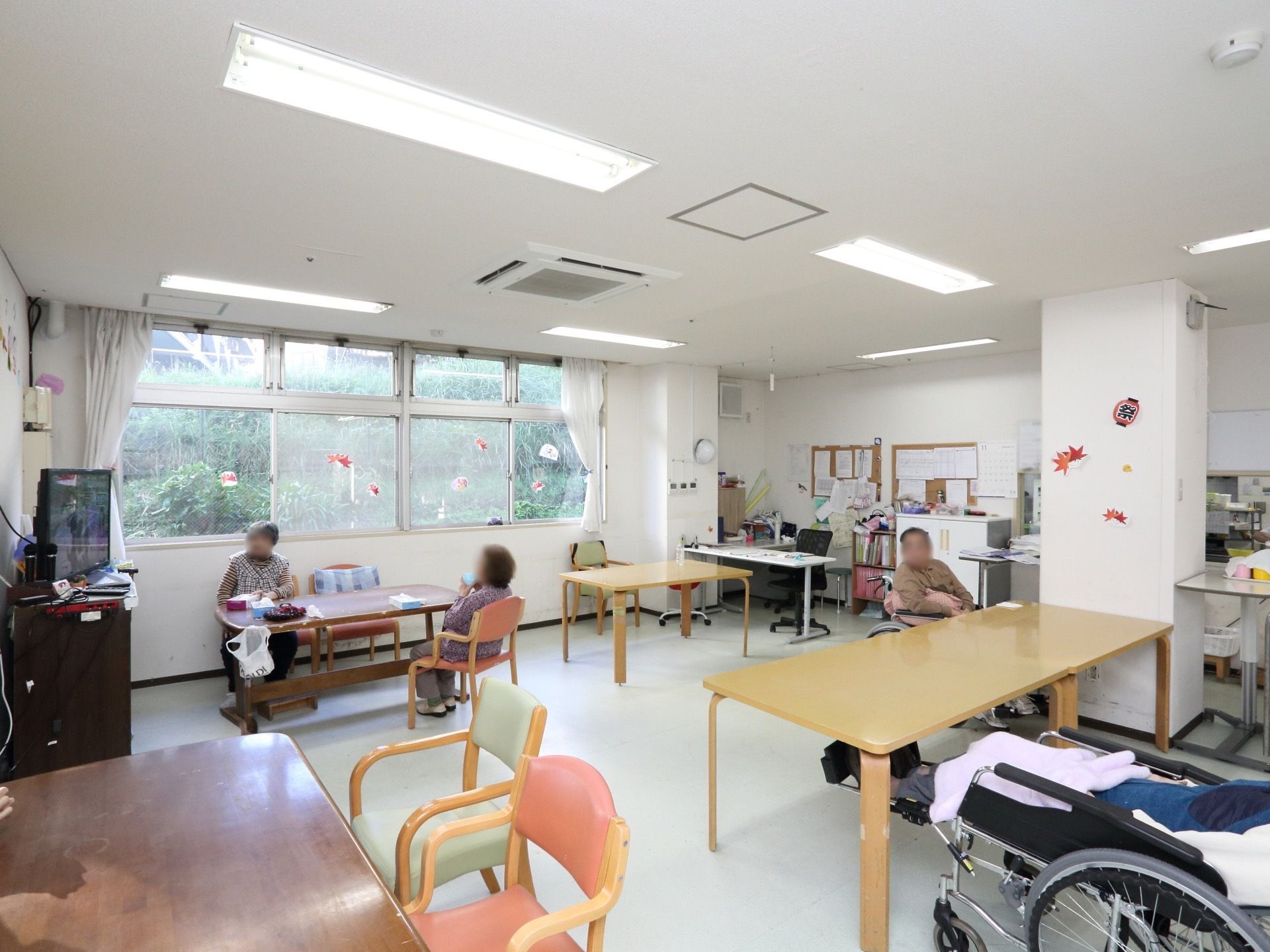 大きなテーブルと椅子が並べられている食堂。壁一面は窓になっており、外には緑が生い茂っている。テレビが置かれている。