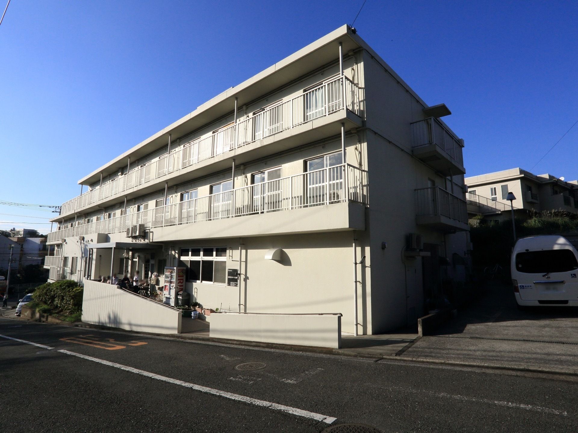 坂道の途中にある、白い大きな建がある。建物のすぐ横は駐車場になっている。玄関へはスロープが続いている。