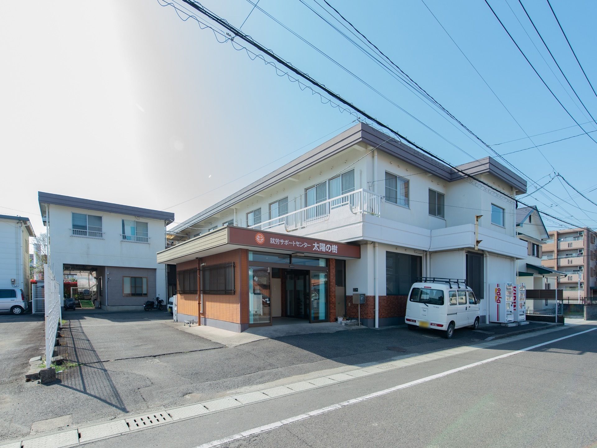 L字型の2階建て施設。敷地前面はアスファルトで舗装されており、通り沿いの自動販売機の近くに自動車が1台駐まっている。