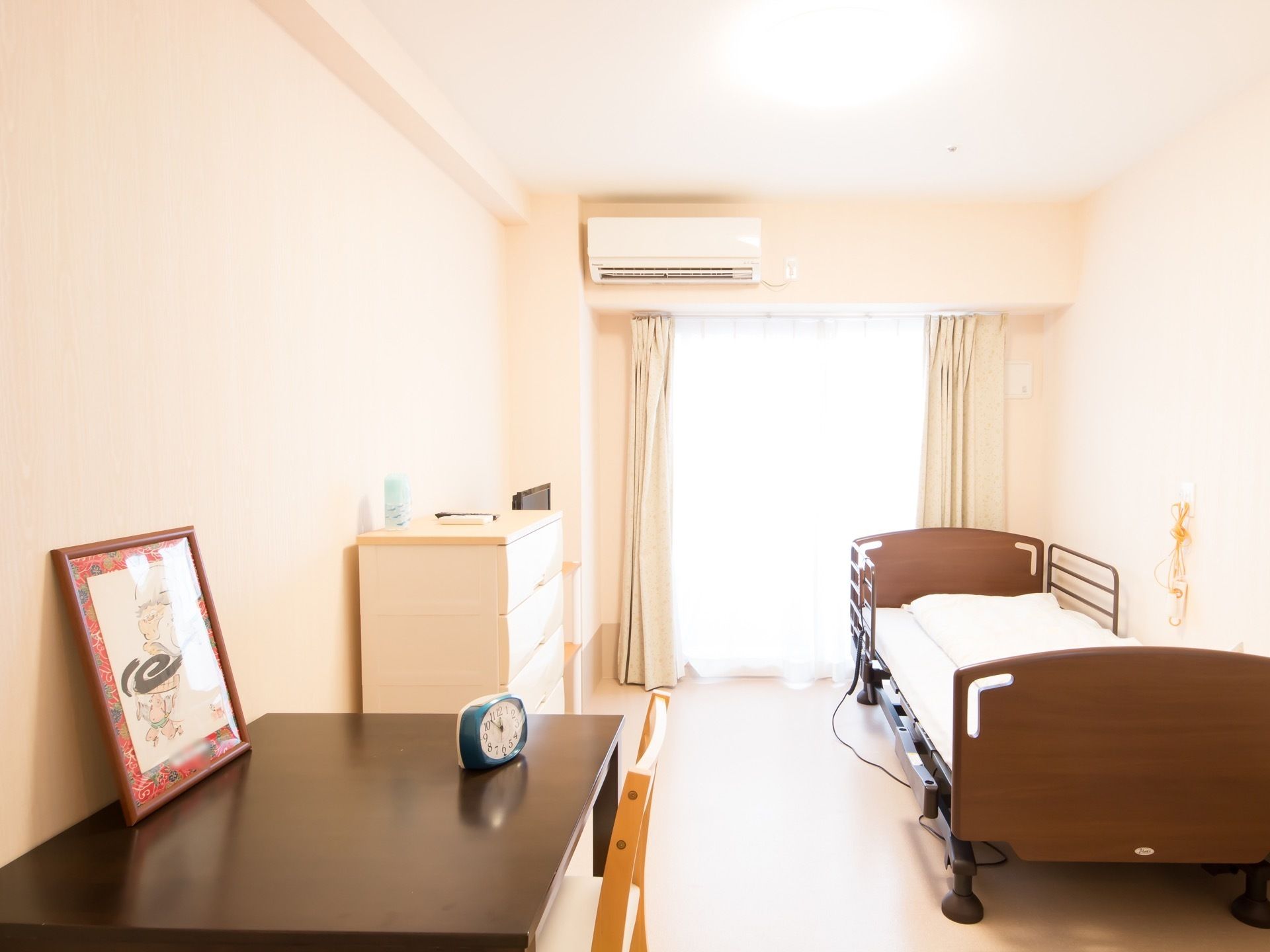 入居者様の居室には、茶色いシングルベッドが用意されている。机の上には、青色の小さな時計が置かれている。